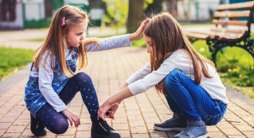 Two young girls sitting on the ground, with one helping the other to tie her shoes